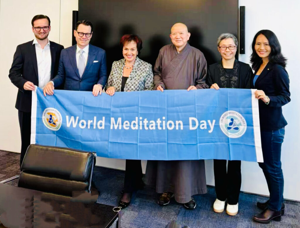 Participants hold a World Meditation Day banner in a meeting room.
